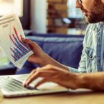 Cropped image of handsome businessman in casual wear using a laptop and examining documents while working in the office