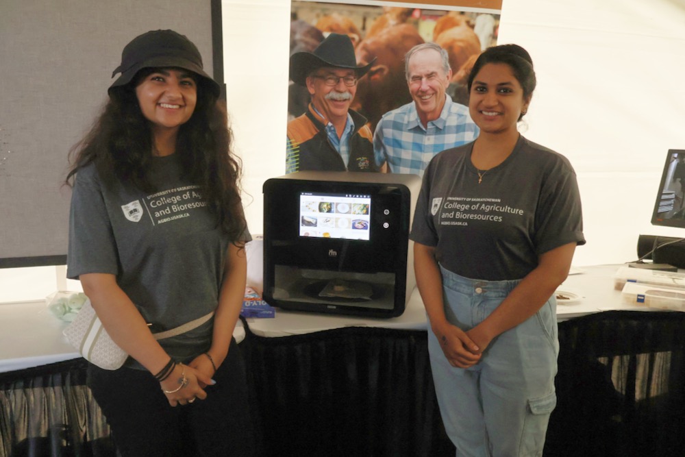 Kashika Sethi (l) and Rhea Thomas Thommana (r) were at Ag in Motion to represent food scientists Drs. Martin Reaney and Michael Nickerson and their 3D printer projects, including their work on printable protein-based materials, such as the pea-based “chicken leg” seen in the machine. (Becky Zimmer photo) 
