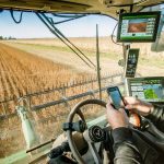 A man sitting in a combine using his smartphone is surrounded by screens and data.