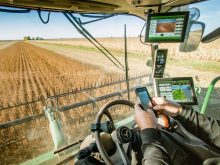 A man sitting in a combine using his smartphone is surrounded by screens and data.
