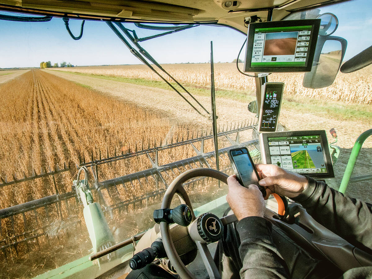 A man sitting in a combine using his smartphone is surrounded by screens and data.