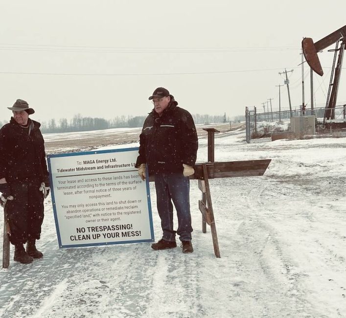 Two men in winter clothing flank wooden barrier with mounted warning sign in snowy field with operating pumpjack in background. Photo: Alexis Kienlen