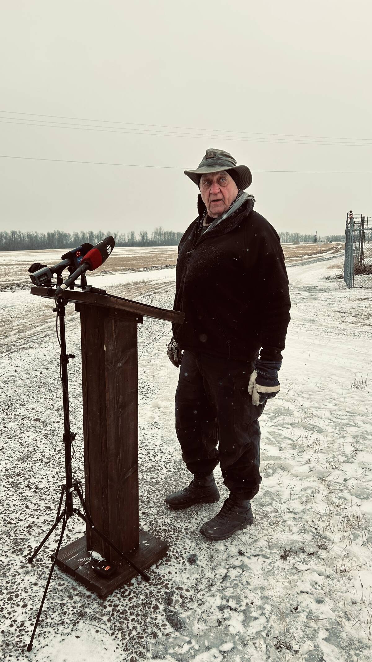 Man at wooden podium with microphones in snowy field with red pumpjack actively operating in background. Photo: Alexis Kienlen