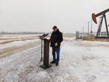Man in cowboy hat and winter coat stands at wooden podium with microphones in snowy rural setting with pumpjack visible in background. Photo: Alexis Kienlen