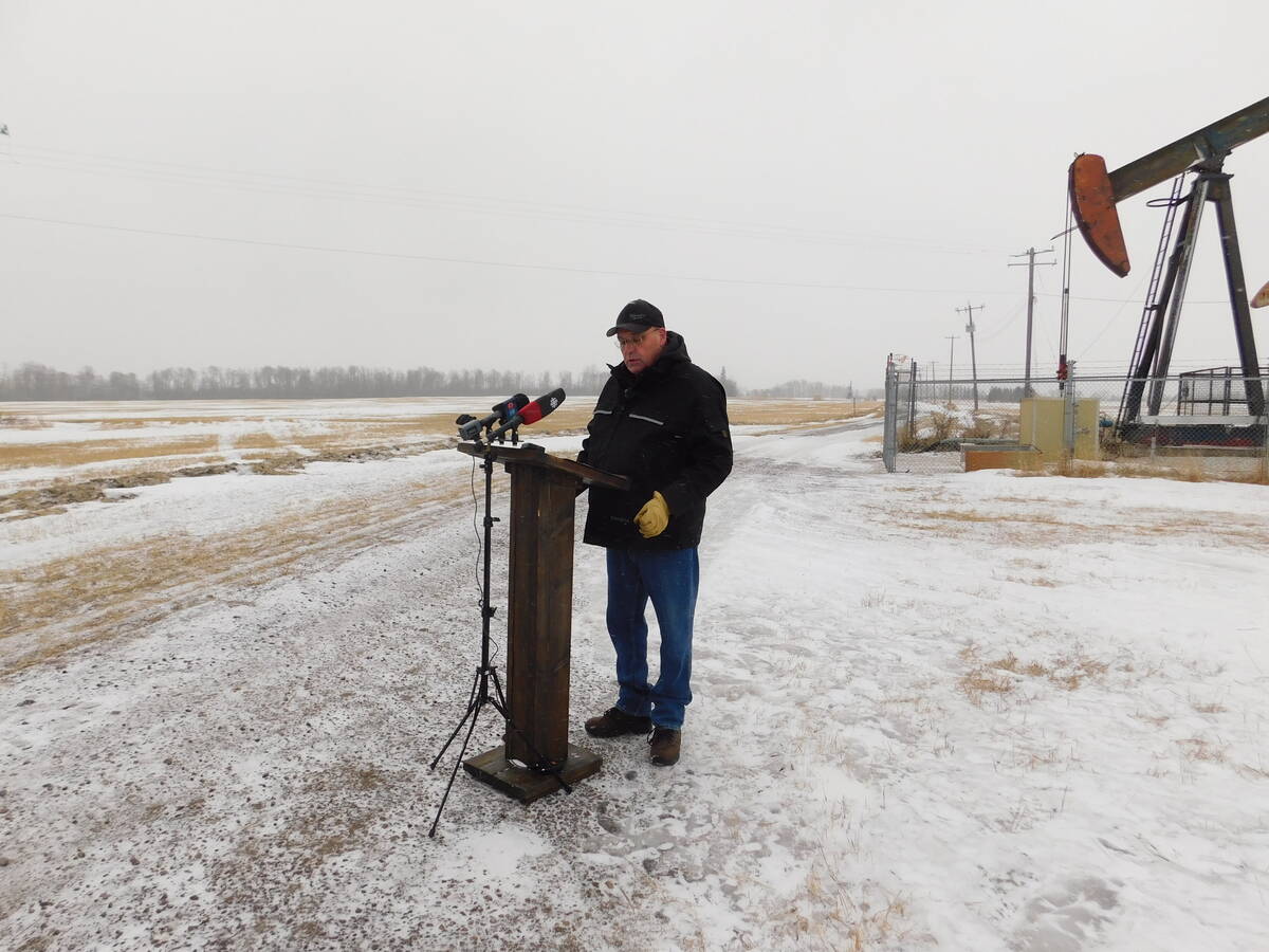 Man in cowboy hat and winter coat stands at wooden podium with microphones in snowy rural setting with pumpjack visible in background. Photo: Alexis Kienlen
