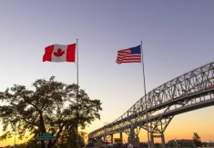 Canadian and American flags flying side by side at sunset with bridge spanning waterway in background. Photo: ehrlif/istock/GettyImages