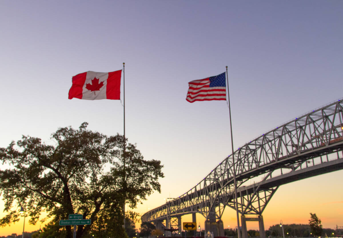 Canadian and American flags flying side by side at sunset with bridge spanning waterway in background. Photo: ehrlif/istock/GettyImages
