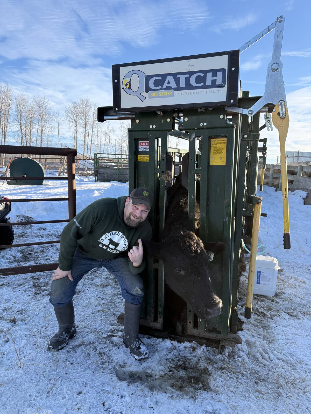Mike Hittinger crouching next to a green Q-Catch squeeze chute with a cow inside, giving a thumbs-up in a snowy farmyard. Photo: Mike Hittinger