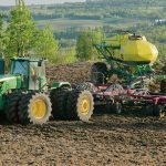 A green tractor pulling a large yellow air seeder across a dark cultivated field, illustrating nitrogen fertilizer application on the Prairies. Photo: Robin Booker