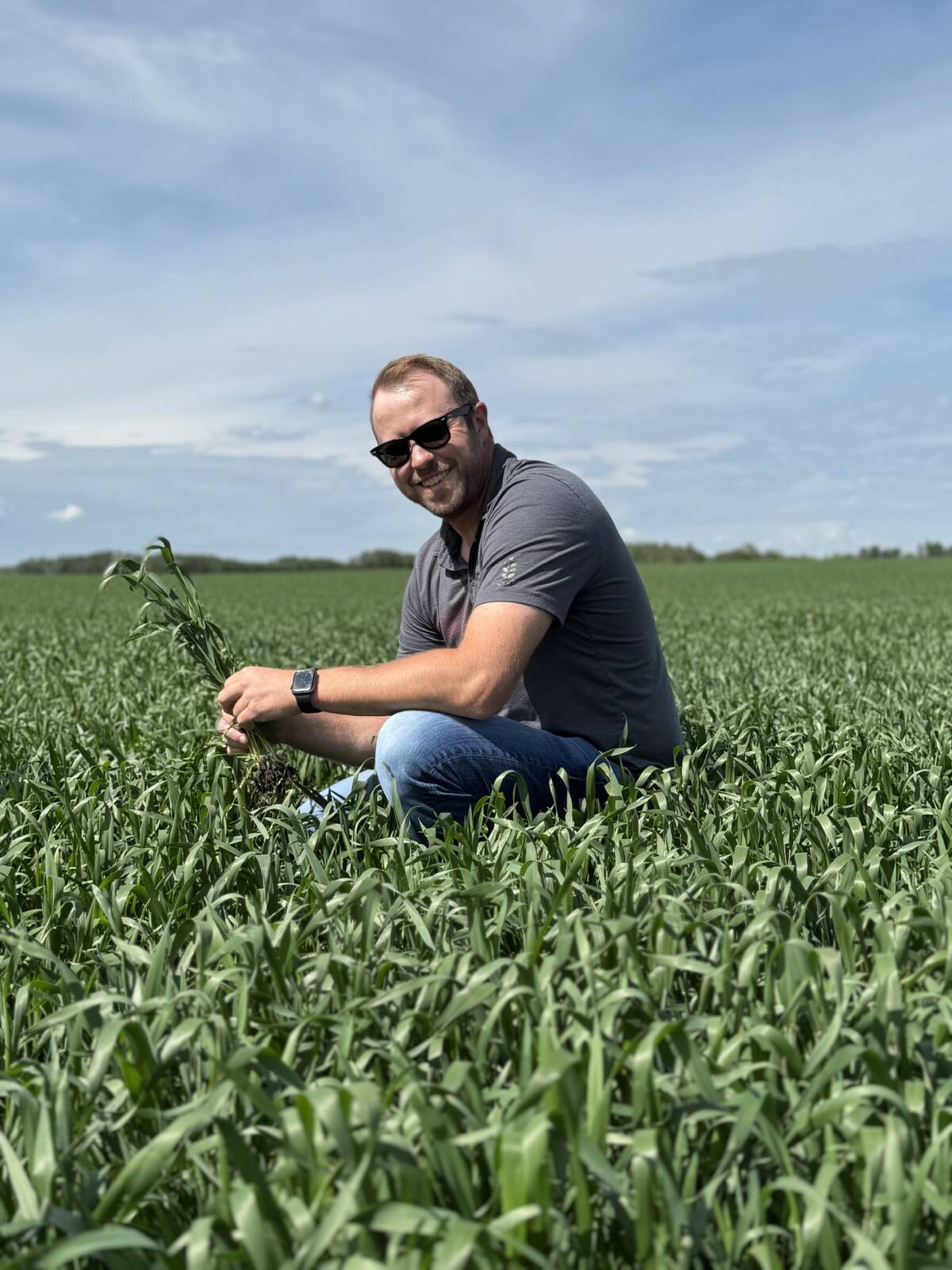 Steve Cowan, an agronomist and producer from near Camrose, Alta., crouching in a green crop field and holding a plant to examine it. Photo: supplied