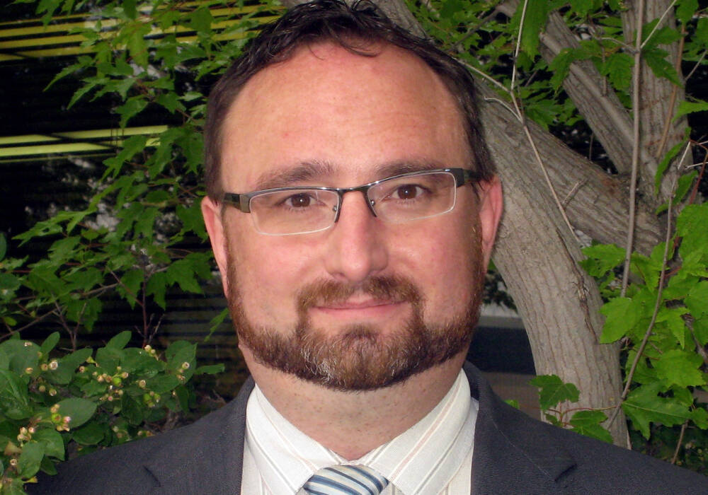 Headshot of Cam Dahl, general manager of Manitoba Pork, wearing glasses and a dark suit jacket, photographed outdoors with green foliage in the background. Photo: file.