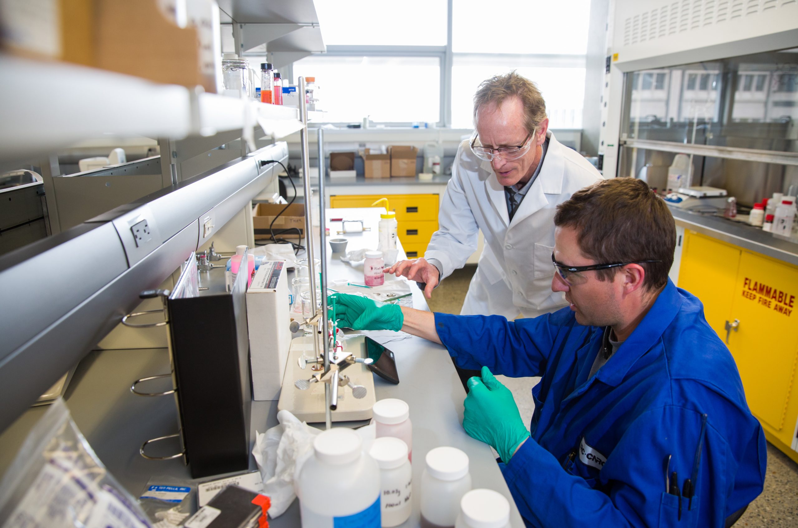 Two researchers in safety glasses working at a laboratory bench, one in a white lab coat and one in a blue lab coat with green gloves, examining samples surrounded by lab equipment and supplies. Photo: Riley Brandt/University of Calgary