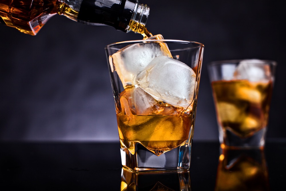 Whisky being poured from a bottle over ice in a glass, with a second glass in the background against a dark backdrop. Photo: Getty Images