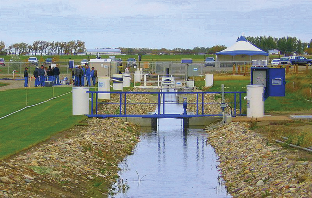 An irrigation canal control structure with blue metal gates and monitoring equipment in southern Alberta, with a group of visitors nearby. Photo: AITC